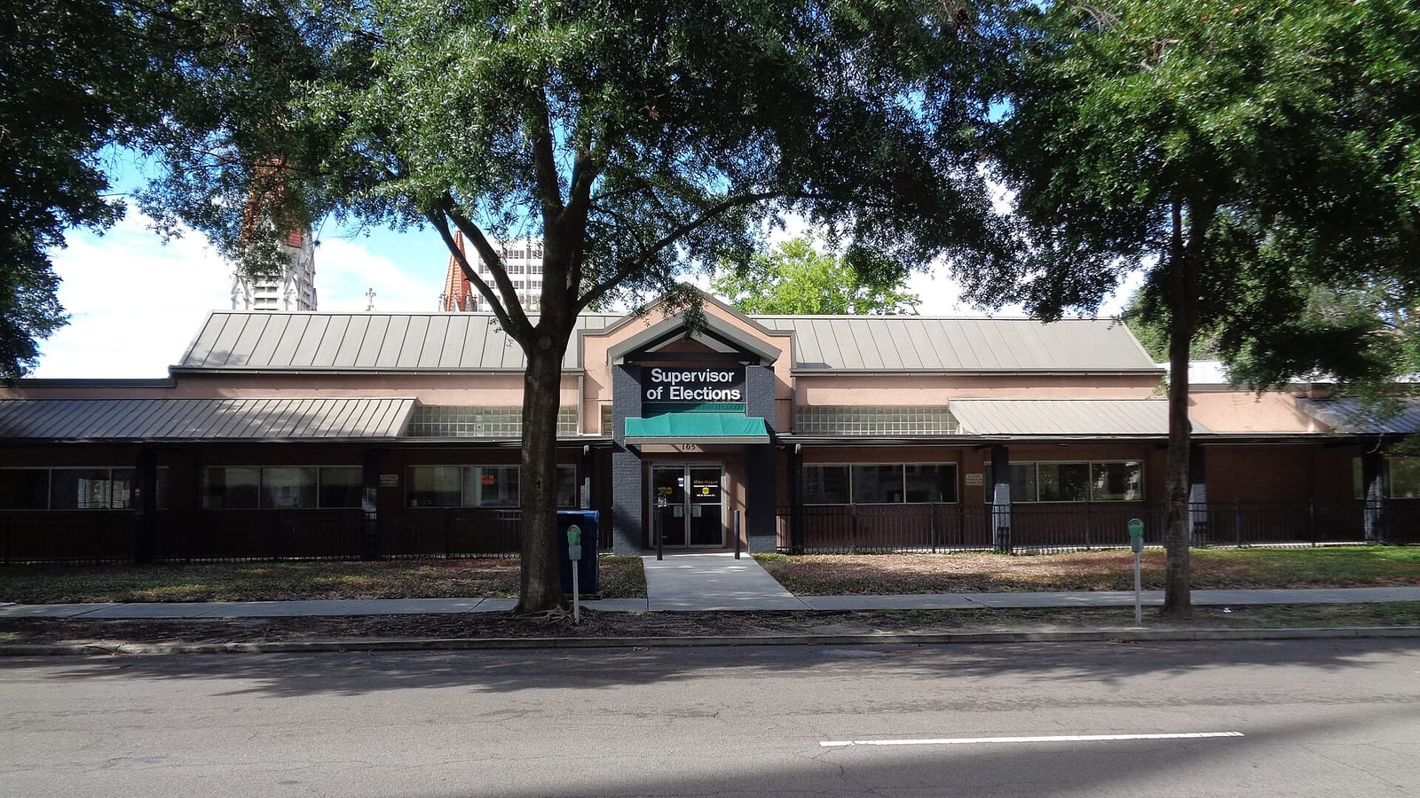 A single-story brick building with a green-roofed entrance under a sign reading Supervisor of Elections, shaded by oak trees beside a street.