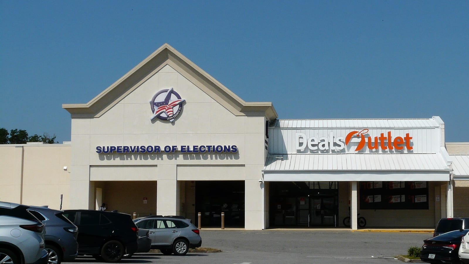 A cream-colored storefront with a peaked gable and a circular star-and-flag emblem above blue letters reading Supervisor of Elections, beside a discount store, with cars parked in front.